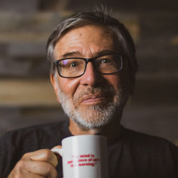 A silver-haired man wearing glasses holds up a coffee mug.